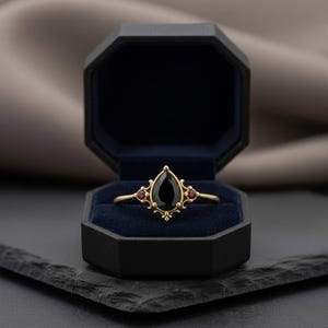 May include: A gold ring featuring a teardrop-shaped black gemstone and two small red stones. The ring is presented in a black, octagonal jewellery box with a blue velvet lining. The ring has an ornate, elegant design.