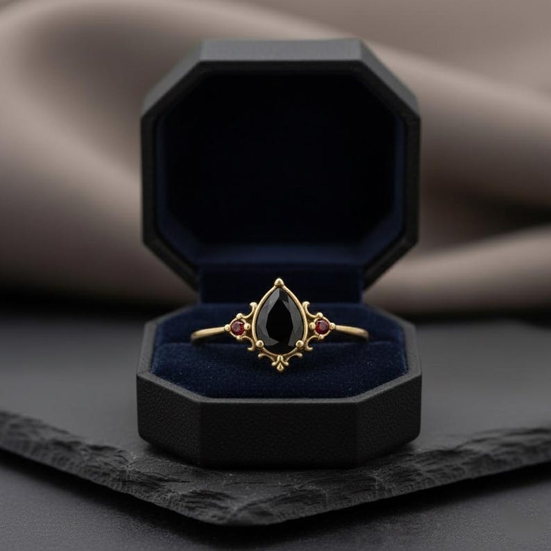 May include: A gold ring featuring a teardrop-shaped black gemstone and two small red stones. The ring is presented in a black, octagonal jewellery box with a blue velvet lining. The ring has an ornate, elegant design.