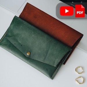 Op de afbeelding: Twee leren portemonnees in groen- en bruintinten. De groene portemonnee heeft een klepsluiting met een messing drukknop. Gouden oorringen staan in de rechter benedenhoek. De portemonnees liggen op een wit oppervlak.