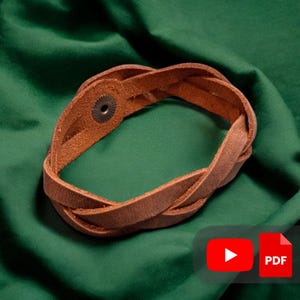 Puede incluir: Una pulsera trenzada de cuero marrón con un cierre de botón a presión de metal. La pulsera está hecha de tiras de cuero entrelazadas, creando una apariencia texturizada y tejida. La pulsera se muestra sobre un fondo de tela verde.