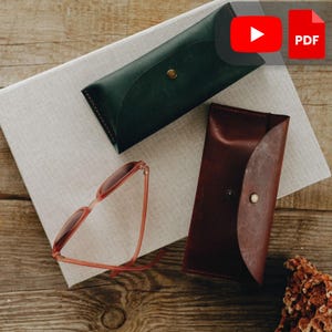 Op de afbeelding: Twee leren brillenkokers in donkergroen en bruin, elk met een drukknoopsluiting. Een paar koraalkleurige zonnebrillen ligt op een neutraal gekleurde stof. De items zijn gerangschikt op een houten oppervlak.
