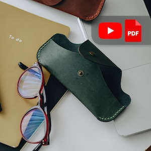 May include: A dark green leather glasses case with a snap closure, beside red-framed glasses. A yellow notebook with the words "To do" is visible. The image also shows a laptop and a brown leather case.