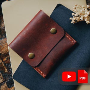 Puede incluir: Una cartera de cuero marrón con una solapa asegurada por dos cierres de botón a presión de color latón. La cartera está encima de un cuaderno beige y otro azul oscuro. Los bordes de la cartera están cosidos con un hilo en contraste.
