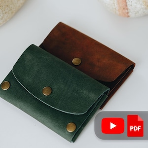 May include: Two leather wallets are shown, one in forest green and the other in brown. Each wallet has a flap closure with a brass snap. The wallets are made of textured leather, with a classic design.