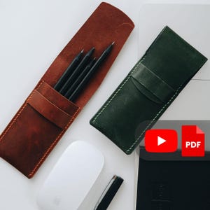 Könnte beinhalten: Zwei Leder-Stifteetuis, eines braun und eines grün, sind geöffnet und zeigen schwarze Stifte. Eine weiße Computermaus und ein Stift sind ebenfalls sichtbar. Das braune Etui enthält vier Stifte, während das grüne Etui leer ist.