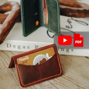 Puede incluir: Un tarjetero de cuero marrón con una tarjeta de crédito dorada visible en el bolsillo delantero. El tarjetero está sobre una superficie de madera, con una cartera de cuero verde en el fondo. El tarjetero está cosido con hilo naranja.