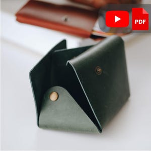 Könnte beinhalten: Ein dunkelgrüner Lederkartenhalter mit einem Messing-Druckknopfverschluss. Das offene Portemonnaie zeigt Innenfächer. Im Hintergrund ist ein weiteres braunes Lederportemonnaie zu sehen. Der Kartenhalter ist für Kredit- und Visitenkarten konzipiert.