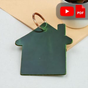 May include: A dark green house-shaped keychain with a copper-colored key ring. The keychain is made of a solid material and has a simple, clean design. The house shape includes a chimney. The background is a light tan color.