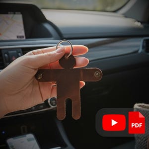 以下が含まれることがあります： 手で持たれた、人の形をした茶色の革製キーホルダー。 キーホルダーには金属製のリングがあり、2つの小さなリベットで固定されています。 背景には車の内装が写っています。