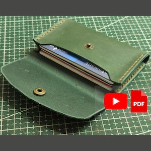 May include: Open, green leather card holder with a brass snap closure. The card holder is filled with multiple cards. The edges are stitched with a contrasting tan thread. The card holder is on a green cutting mat.