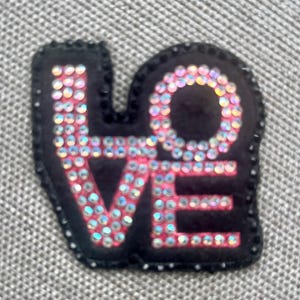 May include: A black patch with the word "LOVE" spelled out in iridescent rhinestones. The patch has a black border with small, black beads. The letters are outlined in pink.