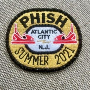 May include: Embroidered patch with the word "PHISH" in black letters on a yellow background. The patch features red roller skates and the text "ATLANTIC CITY N.J. SUMMER 2021". The patch is oval-shaped with a black border.