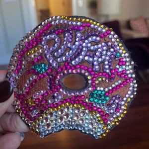 May include: A bedazzled donut-shaped patch with a bite taken out. The patch features a brown base with pink, purple, yellow, and teal rhinestones. The rhinestones are arranged in a pattern that resembles sprinkles and frosting.