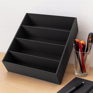 Puede incluir: Un organizador de escritorio negro con cuatro compartimentos angulados. El organizador está hecho de un material mate y se encuentra sobre una superficie de madera de color claro. Un vaso con bolígrafos y herramientas está a la derecha.