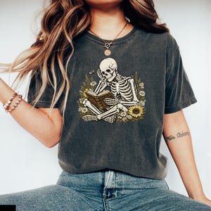 May include: A charcoal grey short-sleeve t-shirt featuring a graphic print of a skeleton sitting cross-legged whilst reading a book, surrounded by yellow sunflowers and green foliage. The shirt has a crew neckline and a relaxed, vintage-washed finish.