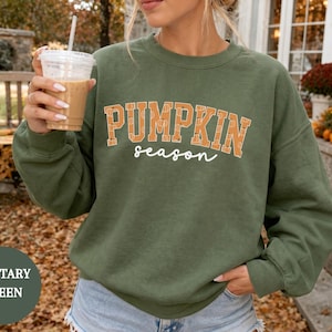 Puede incluir: Una sudadera de cuello redondo color verde militar con el texto PUMPKIN en letras grandes de bloque naranja con un patrón de calabazas, y la palabra season en letra cursiva blanca debajo. La sudadera tiene mangas largas y cuello acanalado.