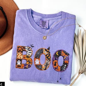 Puede incluir: Una camiseta violeta de manga corta con la palabra BOO impresa en letras grandes. Cada letra está rellena con un patrón de retazos de motivos de Halloween que incluyen fantasmas, calabazas, murciélagos, arañas, esqueletos y dulces de maíz.