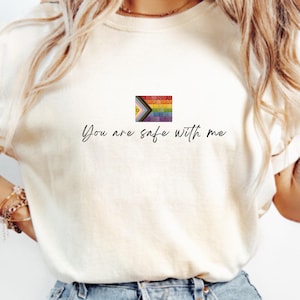Puede incluir: Camiseta de color marfil con un pequeño gráfico de la bandera del Orgullo, multicolor. El texto "You are safe with me" está impreso en una escritura delicada debajo de la bandera. La camiseta tiene mangas cortas y un corte holgado.