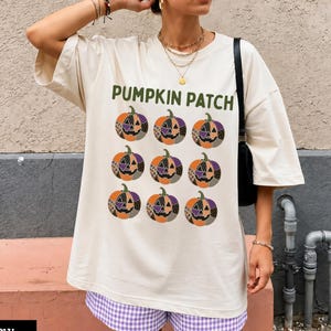 Puede incluir: Una camiseta de manga corta color marfil con el texto PUMPKIN PATCH en letras verdes sobre una cuadrícula de nueve calabazas estampadas en tonos naranja, morado y negro. La camiseta se combina con pantalones cortos de cuadros vichy morados y blancos.