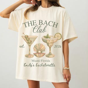 Puede incluir: Camiseta color crema con el texto "THE BACH Club" y "Emily's bachelorette" en verde. La camiseta presenta ilustraciones de cócteles y una concha. El año "2026" también está impreso en la camiseta.