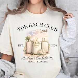 Puede incluir: Camiseta color crema con el texto "THE BACH CLUB" sobre un gráfico de maletas, flores y un avión. El texto "EST. 2026" y "Andrea's Bachelorette MIAMI - FLORIDA" también están impresos en la camiseta.