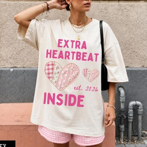 May include: An ivory oversized t-shirt featuring the pink text EXTRA HEARTBEAT INSIDE and est. 2026. The graphic includes two hearts with a patchwork pattern of pink gingham, floral, and polka dot prints, with small white daisy embellishments.