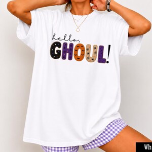 Puede incluir: Una camiseta blanca de manga corta con el texto "hello, GHOUL!" impreso en el pecho. La palabra "hello," está en letra cursiva negra, mientras que las letras de "GHOUL!" están rellenas con varios patrones que incluyen estrellas, puntos y calabazas.