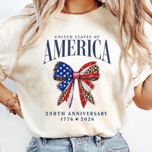 Könnte beinhalten: Ein elfenbeinfarbenes T-Shirt mit dem Text UNITED STATES OF AMERICA in Dunkelblau, einer zentralen Schleifengrafik mit US-Flaggen- und Leopardenmuster sowie dem Text 250TH ANNIVERSARY 1776 Stern 2026 in Dunkelblau unten.