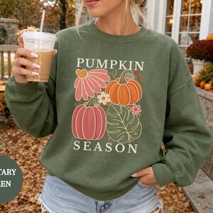 May include: A military green crewneck sweatshirt featuring a graphic print of pumpkins, a pink flower, and a green leaf. The text PUMPKIN SEASON is printed in white above and below the illustration. The sweatshirt has long sleeves and a ribbed collar.