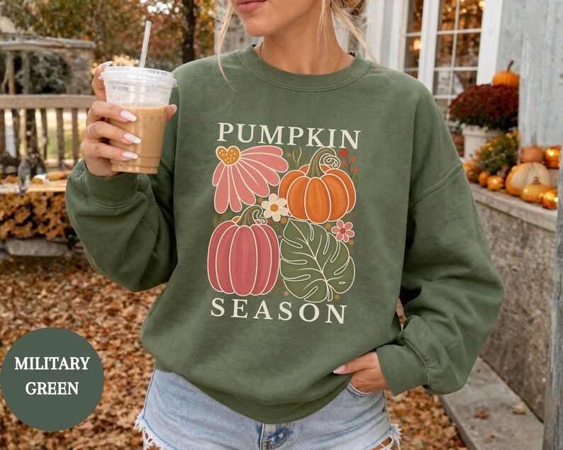 May include: A military green crewneck sweatshirt featuring a graphic print of pumpkins, a pink flower, and a green leaf. The text PUMPKIN SEASON is printed in white above and below the illustration. The sweatshirt has long sleeves and a ribbed collar.
