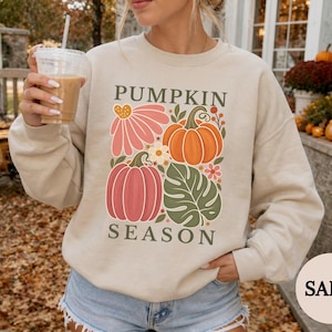 May include: A sand-colored crewneck sweatshirt featuring a graphic print with the text PUMPKIN SEASON. The design includes a pink flower, an orange pumpkin, a red pumpkin, and a green monstera leaf, all arranged in a rectangular composition.