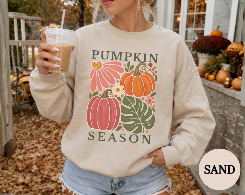 May include: A sand-colored crewneck sweatshirt featuring a graphic print with the text PUMPKIN SEASON. The design includes a pink flower, an orange pumpkin, a red pumpkin, and a green monstera leaf, all arranged in a rectangular composition.