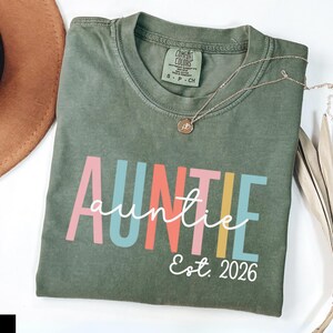 Puede incluir: Una camiseta de manga corta color verde musgo con la palabra AUNTIE en letras mayúsculas grandes y multicolores, con la palabra auntie en letra cursiva blanca superpuesta y el texto Est. 2026 en letra cursiva blanca debajo. Etiqueta Comfort Colors.