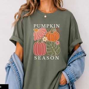 May include: A moss green short-sleeve t-shirt featuring a graphic print with the text PUMPKIN SEASON. The design includes a pink flower, an orange pumpkin, a pink pumpkin, a green monstera leaf, and small floral accents in a grid layout.