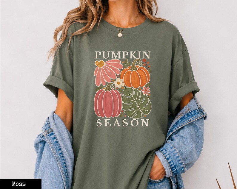 May include: A moss green short-sleeve t-shirt featuring a graphic print with the text PUMPKIN SEASON. The design includes a pink flower, an orange pumpkin, a pink pumpkin, a green monstera leaf, and small floral accents in a grid layout.