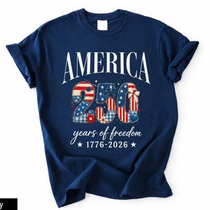 Puede incluir: Una camiseta azul marino de manga corta con el texto blanco AMERICA sobre el número 250, relleno con un patrón patriótico rojo, blanco y azul. Debajo, el texto dice years of freedom 1776-2026 con dos pequeñas estrellas blancas.