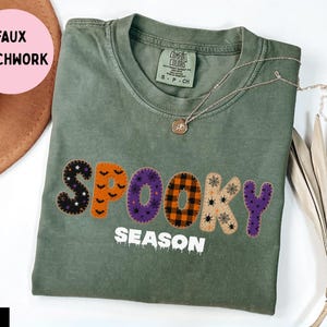 Puede incluir: Una camiseta de manga corta color verde musgo con el texto SPOOKY SEASON en letras de estilo patchwork. Las letras muestran varios patrones que incluyen murciélagos, arañas, telarañas y cuadros en naranja, morado y negro. Tiene cuello redondo.