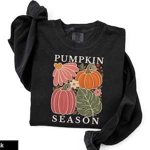 May include: A black long-sleeve shirt featuring a graphic print with the text PUMPKIN SEASON. The design includes two orange pumpkins, a large pink flower, a green monstera leaf, and small floral accents in shades of orange, pink, and white.