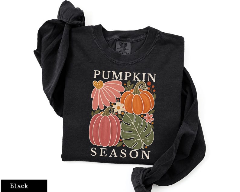 May include: A black long-sleeve shirt featuring a graphic print with the text PUMPKIN SEASON. The design includes two orange pumpkins, a large pink flower, a green monstera leaf, and small floral accents in shades of orange, pink, and white.