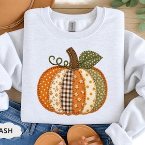Puede incluir: Una sudadera de cuello redondo gris claro con un gráfico central de calabaza compuesto por varios parches de estilo tela. El diseño de la calabaza incluye patrones naranjas, marrones y verdes como lunares, cuadros vichy y estampados florales.