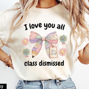 Puede incluir: Camiseta color marfil con el texto "I love you all class dismissed". El diseño incluye un gran lazo, un lápiz, una etiqueta con el nombre "Mrs. Smith" y gráficos florales y de globo en colores pastel.
