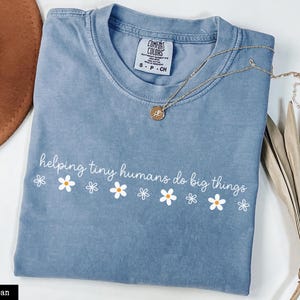 May include: A blue jean coloured t-shirt with a crew neckline and a white graphic print that reads "helping tiny humans do big things" above a row of small white and yellow daisy flowers. The shirt features a Comfort Colors brand neck label.