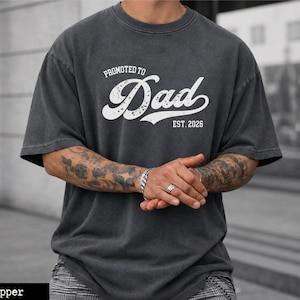 May include: Dark gray t-shirt with the text "Promoted to Dad EST. 2026" in white script. The shirt has a vintage, worn look. The model is wearing a silver bracelet and rings.