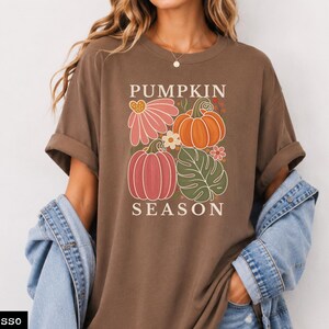 May include: An espresso brown short-sleeve t-shirt featuring a graphic print with the text PUMPKIN SEASON. The design includes two pumpkins, a large pink flower, a small white flower, and a green monstera leaf in a block-style arrangement.