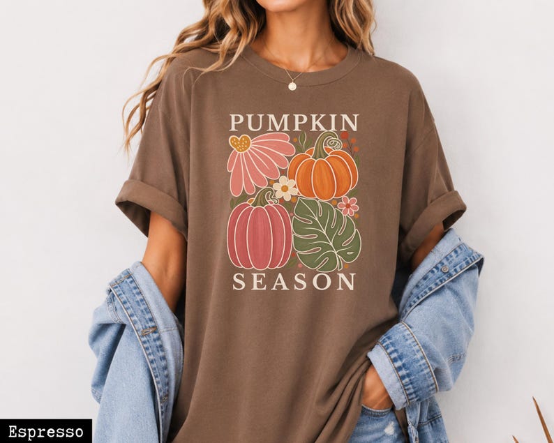May include: An espresso brown short-sleeve t-shirt featuring a graphic print with the text PUMPKIN SEASON. The design includes two pumpkins, a large pink flower, a small white flower, and a green monstera leaf in a block-style arrangement.