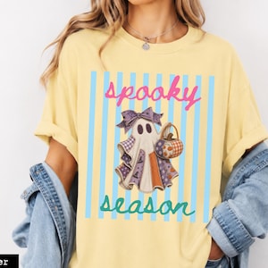 Puede incluir: Una camiseta de color amarillo mantequilla con un gráfico de un fantasma que lleva un lazo morado estampado y sostiene una cesta de calabaza. El diseño incluye rayas verticales azul claro y el texto "spooky season" en letra cursiva rosa y verde azulado.