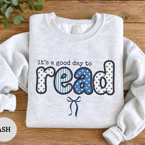 Puede incluir: Una sudadera gris ceniza de cuello redondo con el texto "it's a good day to read" en patrones azules y blancos. La palabra "read" aparece en letras grandes y negritas con diseños de puntos y rayas, acentuada por un pequeño lazo azul.