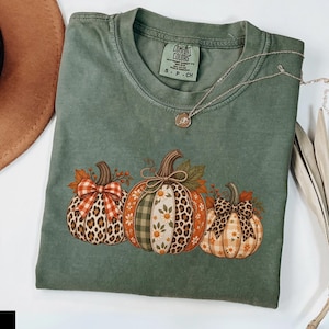 Puede incluir: Una camiseta de manga corta color verde musgo con un gráfico de tres calabazas con patrones variados que incluyen estampado de leopardo, cuadros y flores. Las calabazas están decoradas con lazos y hojas de otoño. Incluye etiqueta de Comfort Colors.