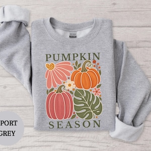 May include: A sport grey crewneck sweatshirt featuring a graphic print of two pumpkins, a large pink flower, and a green monstera leaf. The text PUMPKIN SEASON is printed in dark green above and below the illustration on the front of the garment.
