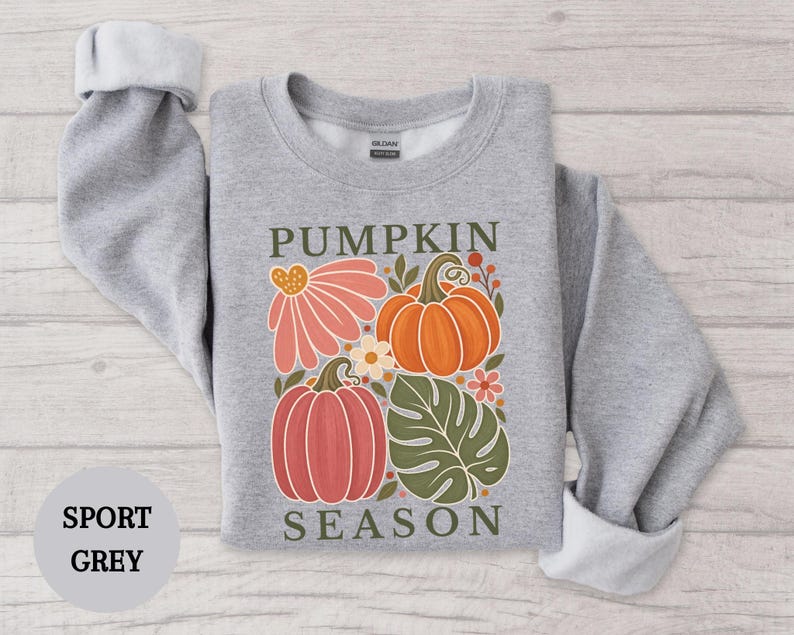 May include: A sport grey crewneck sweatshirt featuring a graphic print of two pumpkins, a large pink flower, and a green monstera leaf. The text PUMPKIN SEASON is printed in dark green above and below the illustration on the front of the garment.